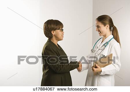 Stock Photo - Caucasian mid-adult female doctor shaking hands with African American middle-aged female patient.. Fotosearch