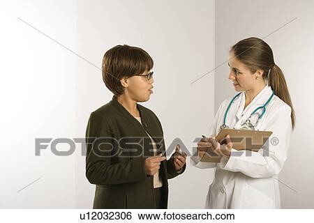 Stock Photograph - Caucasian mid-adult female doctor talking with African American middle-aged female patient.. Fotosearch