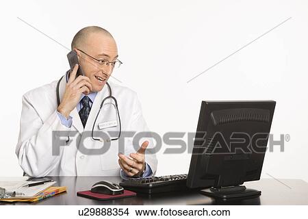 Picture - Caucasian mid adult male physician sitting at desk with laptop computer talking on cellphone.. Fotosearch