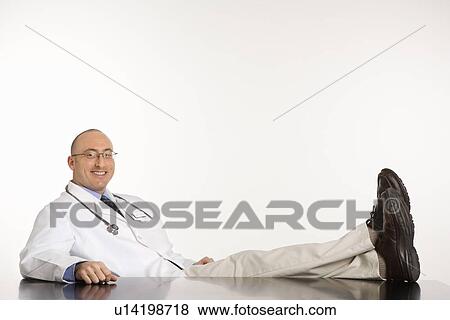 Stock Photo - Caucasian mid adult male physician sitting with feet on desk smiling.. Fotosearch