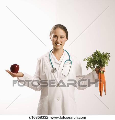 Stock Image - Portrait of Caucasian mid-adult female doctor holding red apple and bunch of carrots smiling and looking at viewer.. Fotosearch