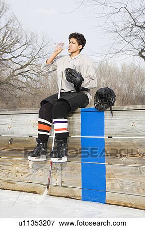 Stock Photo - Boy in ice hockey uniform holding hockey stick sitting on sidelines drinking water.. Fotosearch