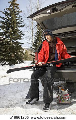 Stock Photography - Caucasian male teenager sitting on back of SUV holding skis.. Fotosearch