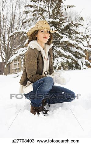 Stock Image - Caucasian young adult female looking at viewer while kneeling in snow with snowball and wearing straw cowboy hat.. Fotosearch