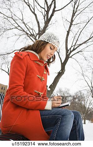 Picture - Low angle view of Caucasian young adult female in winter clothing sitting on park bench using PDA.. Fotosearch