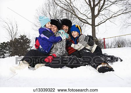 Stock Image - Portrait of happy Caucasian family of four sitting in snow smiling at veiwer.. Fotosearch