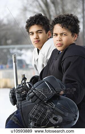 Stock Photo - Two boys in ice hockey uniforms sitting on ice rink sidelines looking. Fotosearch