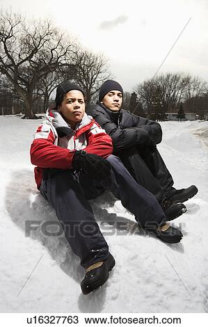 Stock Image - Two boys sitting in snow wearing coats and hats.. Fotosearch