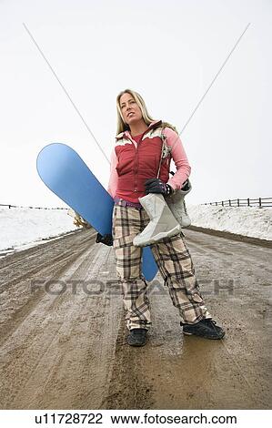 Stock Image - Woman in winter clothes standing holding snowboard and boots. Fotosearch