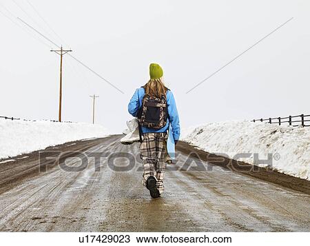 Stock Image - Young woman in winter clothes standing on muddy dirt road holding snowboard and boots.. Fotosearch
