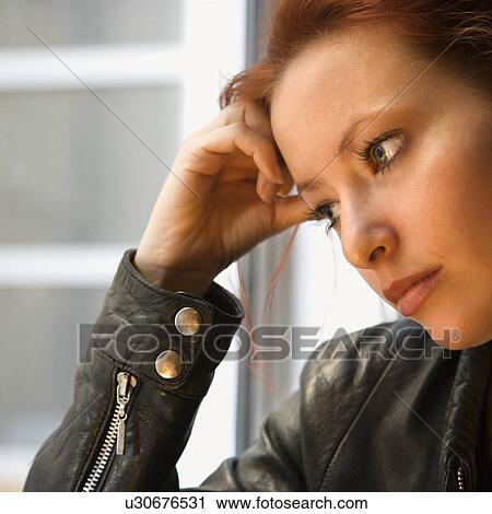 Stock Image - Pretty redhead young woman indoors holding head on hand looking out window. Fotosearch