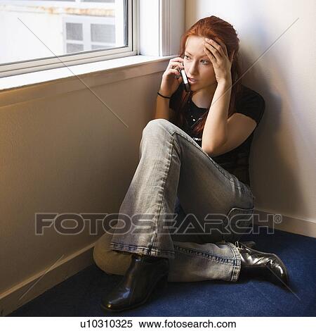 Stock Photography - Pretty redhead young woman sitting on floor indoors by window talking on cellphone.. Fotosearch