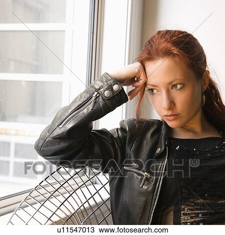 Stock Image - Pretty redhead young woman sitting indoors on metal chair looking out window.. Fotosearch