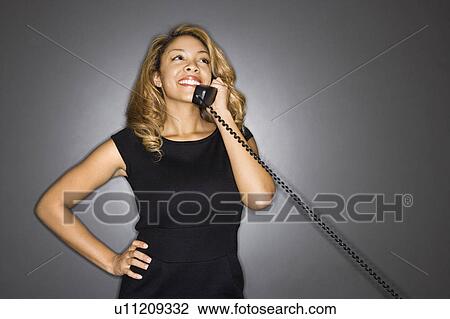 Stock Image - Pretty woman holding telephone receiver to ear smiling standing against gray background.. Fotosearch