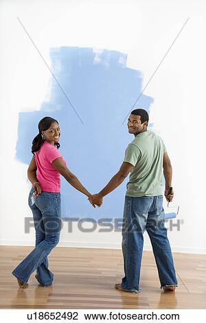 Stock Image - African American couple standing together in front of half-painted wall looking at viewer.. Fotosearch