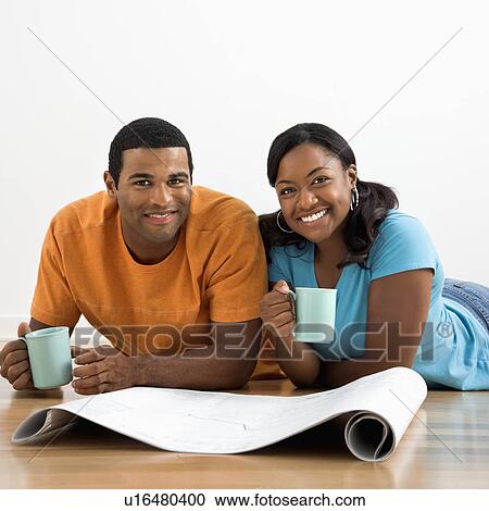 Stock Image - African American male and female couple with architectural blueprints drinking coffee.. Fotosearch