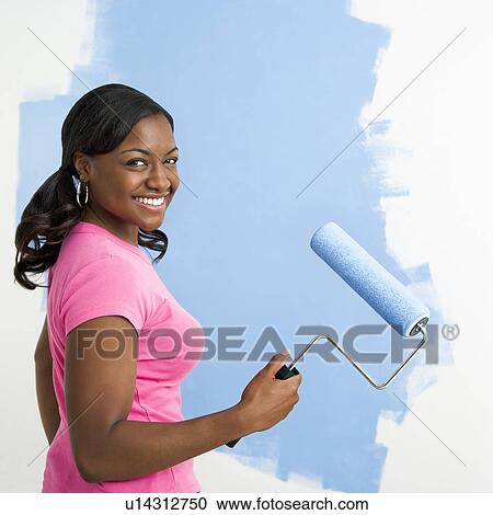 Stock Image - African American woman painting wall blue smiling at viewer.. Fotosearch