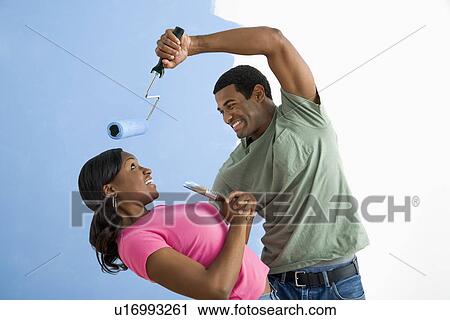 Stock Image - African American young man pretending to paint woman's face with paint roller.. Fotosearch