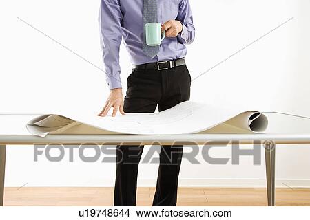Business man standing with coffee cup next to table with blueprints View Large Photo Image Picture - Business man standing with coffee cup next to table with blueprints. Fotosearch