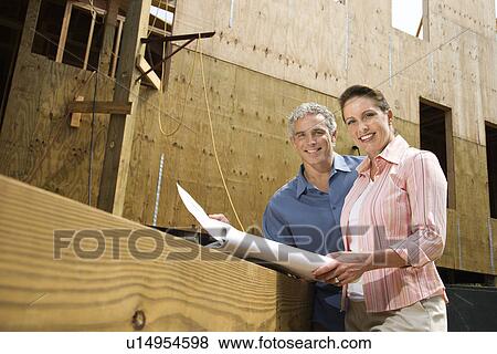 Stock Photo - Caucasian mid-adult male and female holding blueprints looking at viewer.. Fotosearch