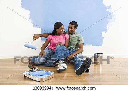 Stock Image - Couple relaxing together next to half-painted wall and painting supplies. Fotosearch