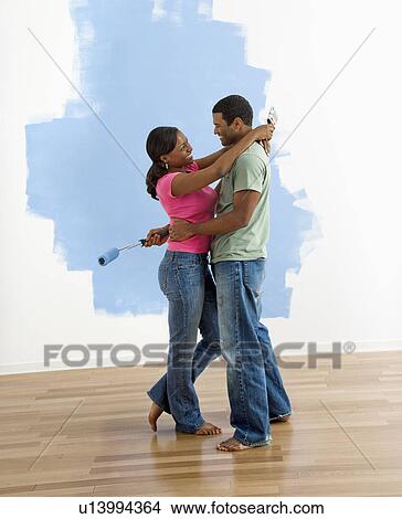 Picture - Couple standing together hugging in front of half-painted wall. Fotosearch