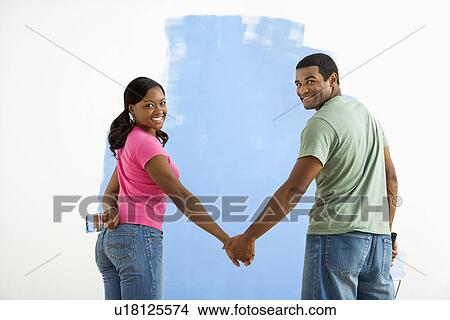 Picture - Couple standing together in front of half-painted wall looking at viewer. Fotosearch