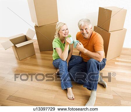 Stock Photo - Middle-aged couple sitting on floor among cardboard moving boxes with coffee.. Fotosearch