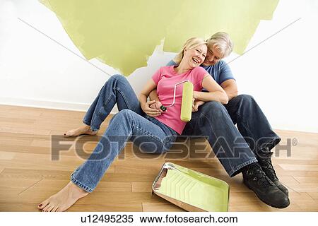 Stock Photography - Portrait of happy adult couple sitting in front of half-painted wall with paint supplies laughing. Fotosearch