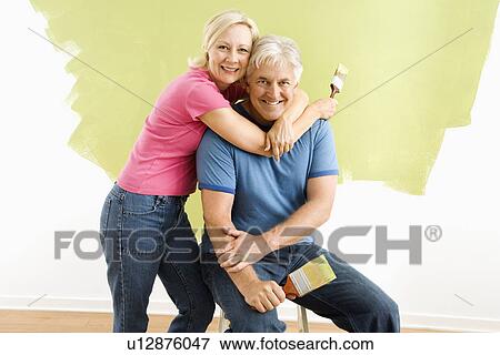 Stock Photo - Portrait of smiling adult couple sitting in front of half-painted wall with paintbrushes. Fotosearch
