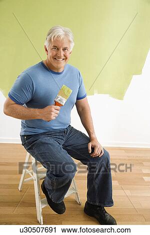 Stock Image - Portrait of smiling adult man sitting in front of half-painted wall with paintbrush.. Fotosearch