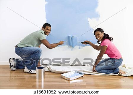Stock Image - Smiling African American male and female couple painting wall blue.. Fotosearch