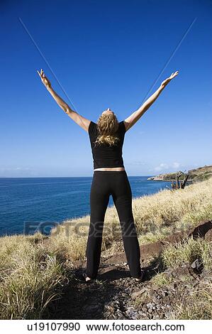 Stock Image - Back view of mid-adult woman standing on Maui, Hawaii coast with arms outstretched and head thrown back. Fotosearch
