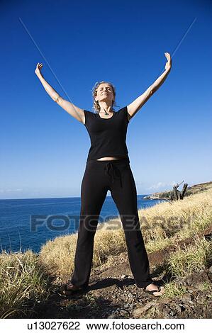 Stock Image - Caucasian mid-adult woman standing on Maui, Hawaii coast with arms outstretched and eyes closed.. Fotosearch