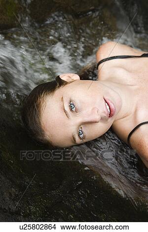 Picture - Caucasian young adult woman laying in freshwater stream looking at viewer.. Fotosearch