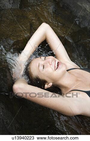 Stock Image - Caucasian young adult woman laying in freshwater stream laughing with arms over head.. Fotosearch