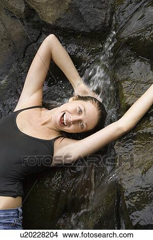 Picture - Caucasian young adult woman standing under small fresh waterfall laughing with arms over head and looking at viewer.. Fotosearch