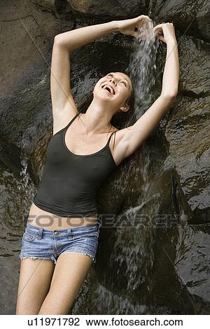 Stock Image - Caucasian young adult woman standing under small fresh waterfall laughing with arms over head.. Fotosearch