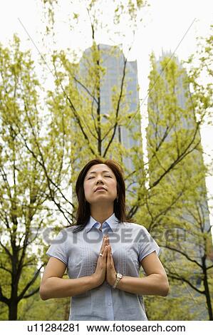 Stock Image - Mid-adult Asian woman standing outside with trees and skyscrapers behind her meditating.. Fotosearch