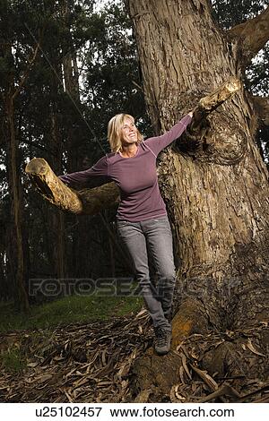 Stock Photo - Middle-aged Caucasian woman smiling standing holding tree in forest.. Fotosearch
