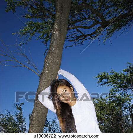 Stock Image - Young adult Asian female holding a tree and looking at viewer.. Fotosearch