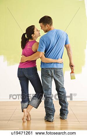 Attractive couple standing in front of partially painted wall with arms around each other smiling View Large Photo Image Stock Image - Attractive couple standing in front of partially painted wall with arms around each other smiling. Fotosearch