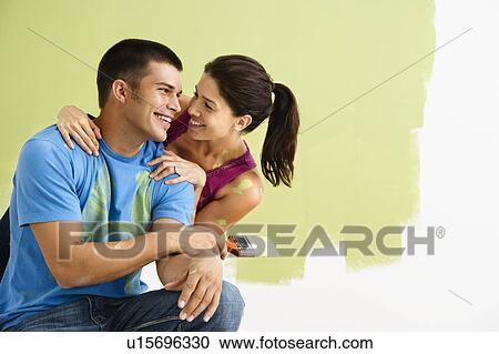 Stock Image - Couple smiling and laughing while painting interior wall of home.. Fotosearch