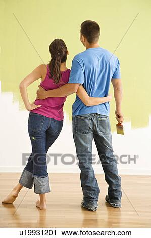 Stock Image - Couple standing in front of partially painted wall with arms around each other.. Fotosearch