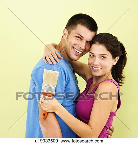 Stock Photo - Happy smiling couple holding paintbrush in front of freshly painted interior wall.. Fotosearch