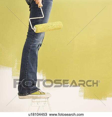 Stock Image - Legs of woman standing on stepladder holding paint roller with painted wall.. Fotosearch