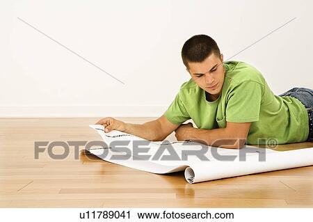 Stock Image - Man lying on floor in home reading house plans. Fotosearch