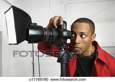 Stock Image - African American young male adult looking through camera on tripod.. Fotosearch