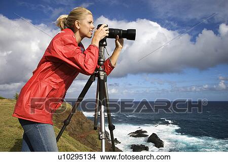 Picture - Woman looking through camera on tripod on cliff overlooking ocean in Maui, Hawaii. Fotosearch
