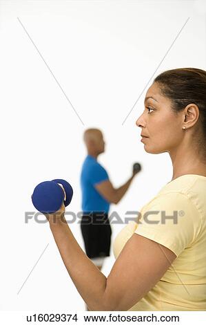 Picture - Close up of smiling mid adult woman exercising using dumbbells with mid adult man in background.. Fotosearch